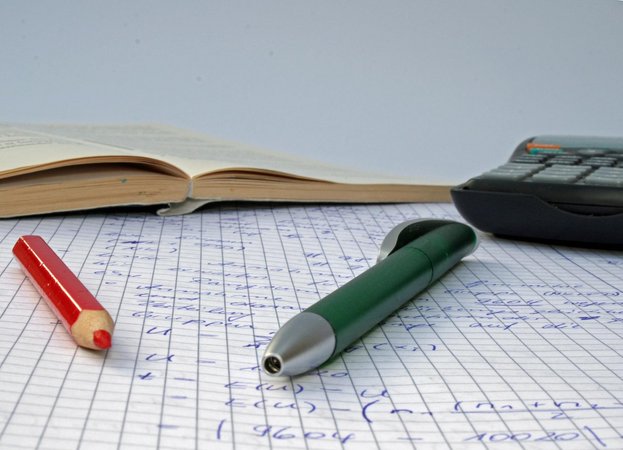 Stifte, aufgeschlagenes Buch und Taschenrechner auf beschriebenem kariertem Blatt Papier